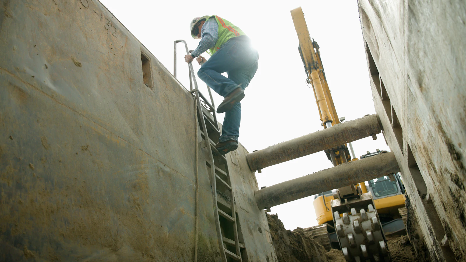 Crew member climbing a ladder out of a shored utility trench, excavator working behind
